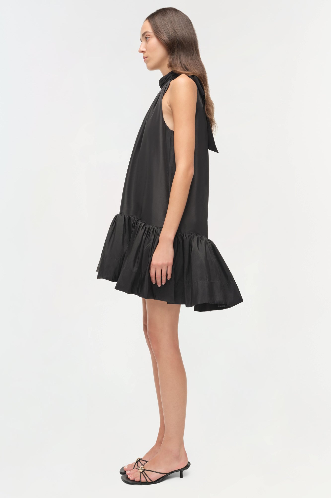 Black sleeveless mini dress with bow neckline and tiered ruffle hem, shown on model in side profile.