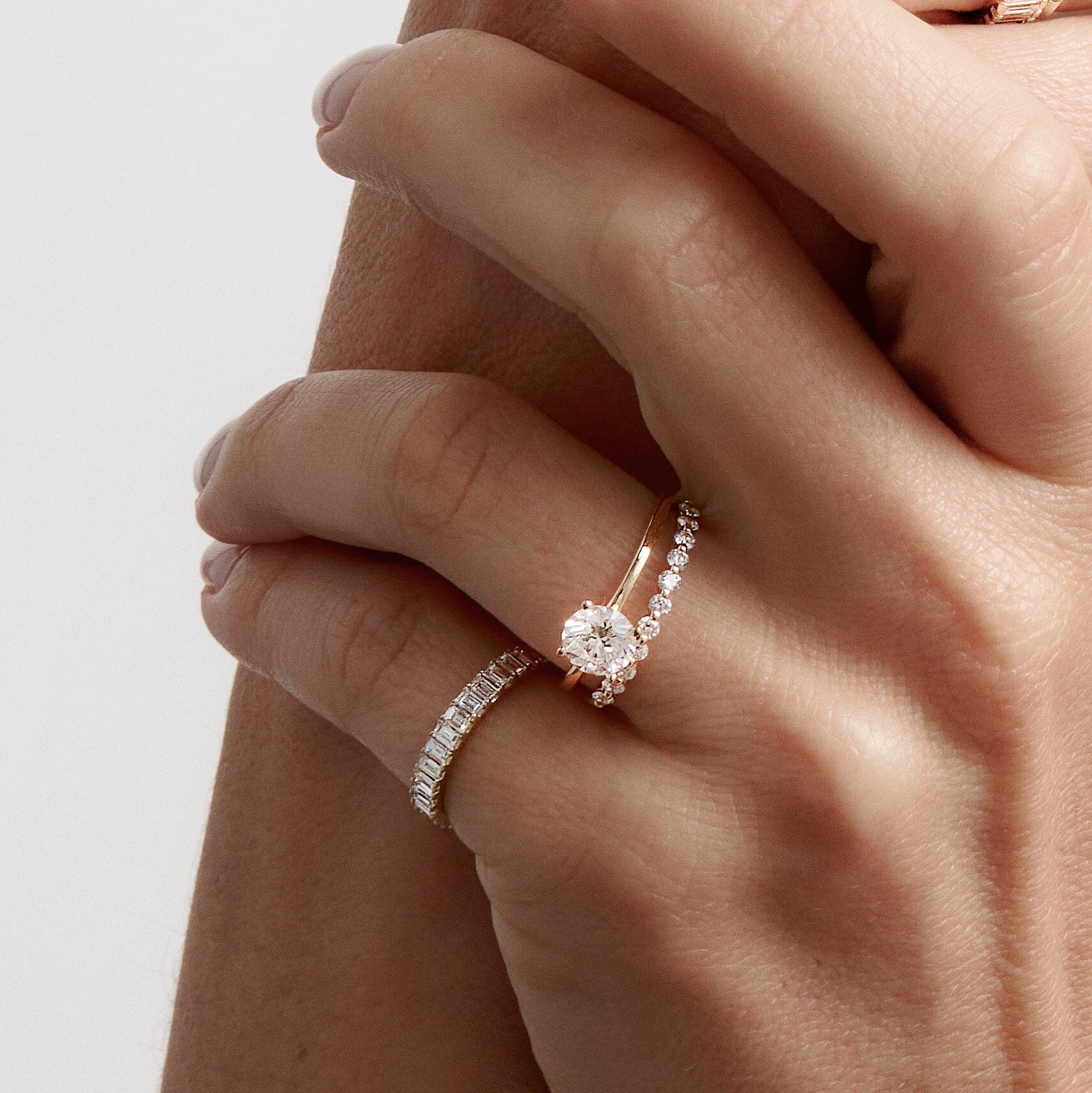Lifestyle Model Close-up of a woman's gently folded hand showcasing three gold and diamond rings against a light background. On the lower finger, she wears a single delicate prong-set diamond band. On the adjacent higher finger, she wears a stacked pair featuring a round diamond solitaire ring paired with another matching prong-set diamond band.