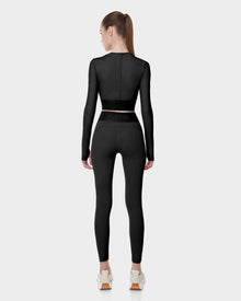 A woman with a ponytail faces away, showcasing athletic style in the Rudsak L523016T DOREEN - BLACK womens long sleeve sports top and matching leggings with branded waistband, paired with white sneakers, against a plain light gray background.