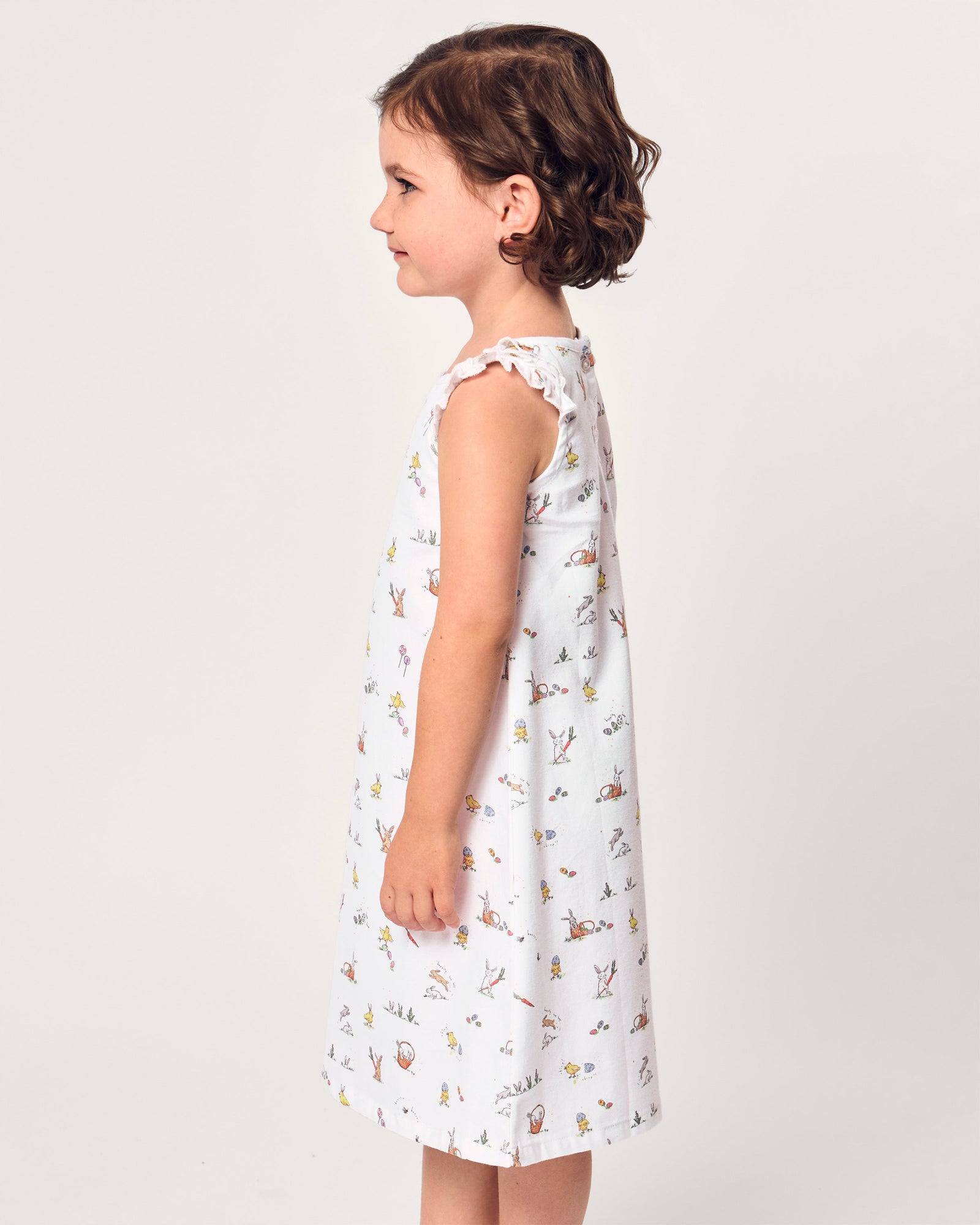 A young girl with short brown hair smiles in the Girl's Twill Amelie Nightgown in Easter Frolic, a white sleeveless dress featuring playful animal and nature prints, standing against a plain light background.