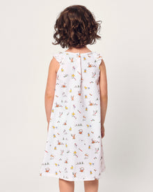 A young girl with short brown hair smiles in the Girl's Twill Amelie Nightgown in Easter Frolic, a white sleeveless dress featuring playful animal and nature prints, standing against a plain light background.