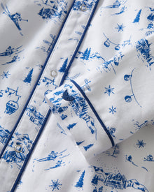 The Baby's Twill Cambridge Romper in Chalet Toile is a white long-sleeve, full-length onesie with blue alpine scene prints and blue piping on the collar, cuffs, and front placket.