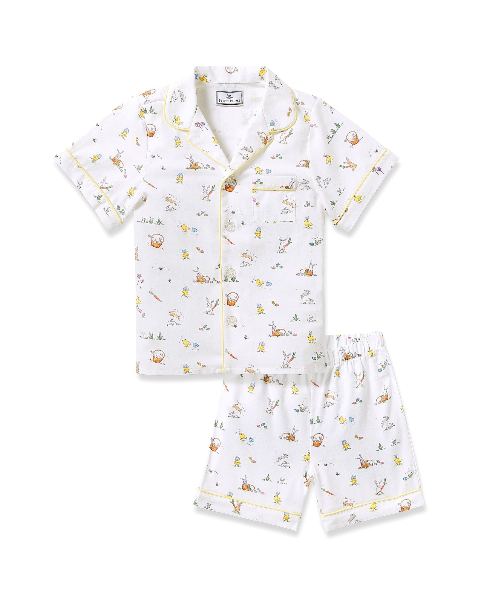 Two smiling children in luxurious Petite Plume sleepwear sit on the floor. They sport matching Kids Twill Short Sets in an Easter Frolic print. One child cuddles a stuffed bunny, embodying comfort and joy against a plain backdrop.