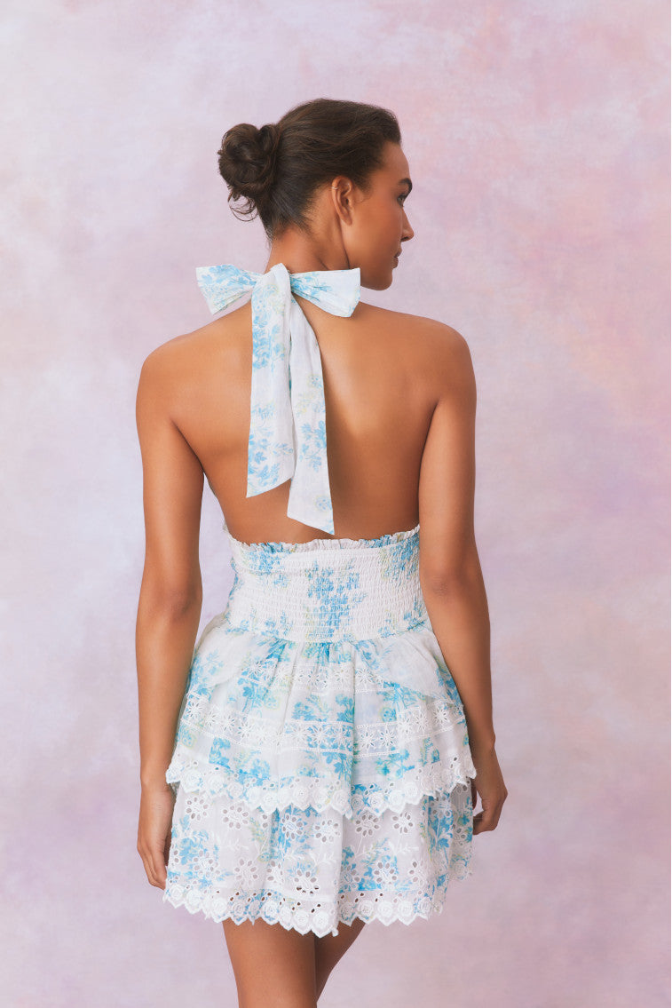White halter dress with a blue floral pattern, halter ties in bow at neck and below, is a wide smocked waistband and the skirt has two ruffled tiers.