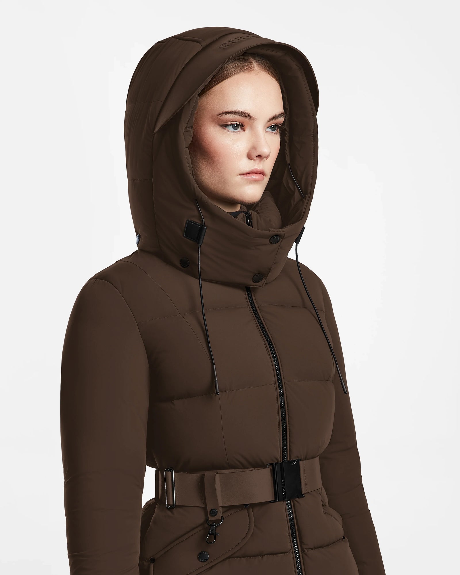 A woman models the Rudsak ADELINE - 8124544 Mocha brown quilted short down puffer jacket with a belt, standing against a white background and gazing slightly to the side, highlighting this stylish womens winter coat.