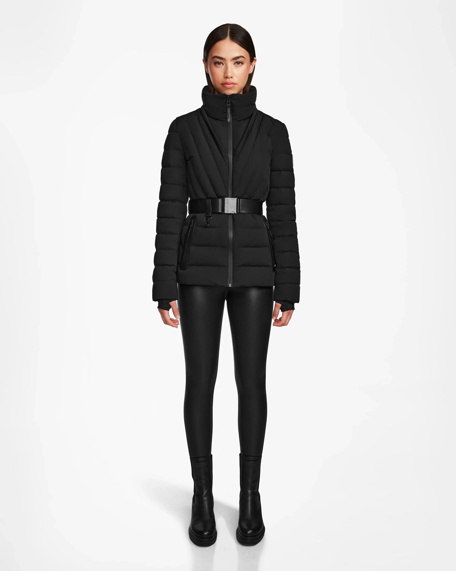 A woman faces forward against a white background, wearing the Rudsak ALISA - L124506 Black quilted down puffer jacket with a vegan leather belt, paired with black shiny leggings and black ankle boots.