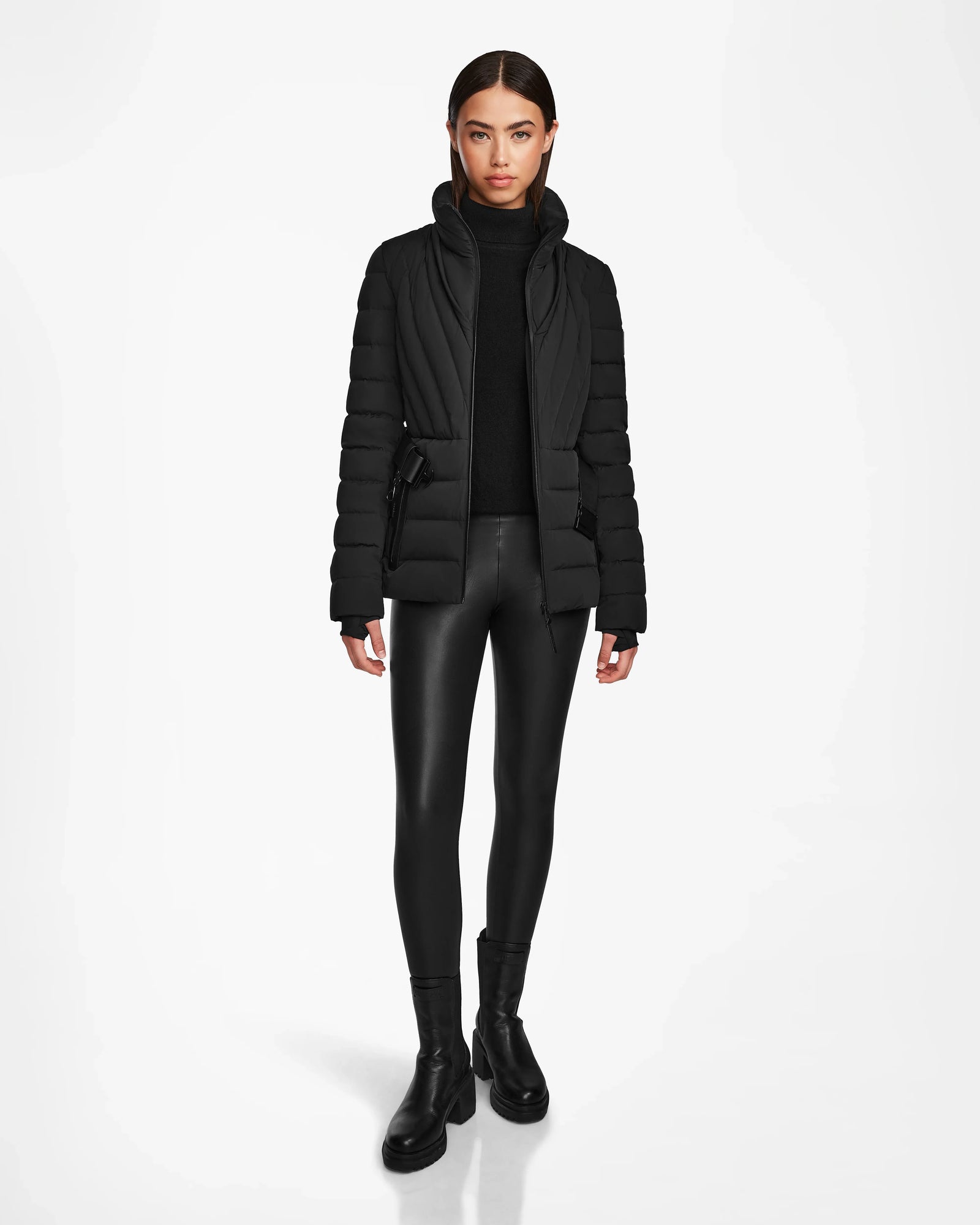 A woman faces forward against a white background, wearing the Rudsak ALISA - L124506 Black womens quilted down puffer jacket, paired with a black turtleneck sweater, faux leather leggings, and black boots.