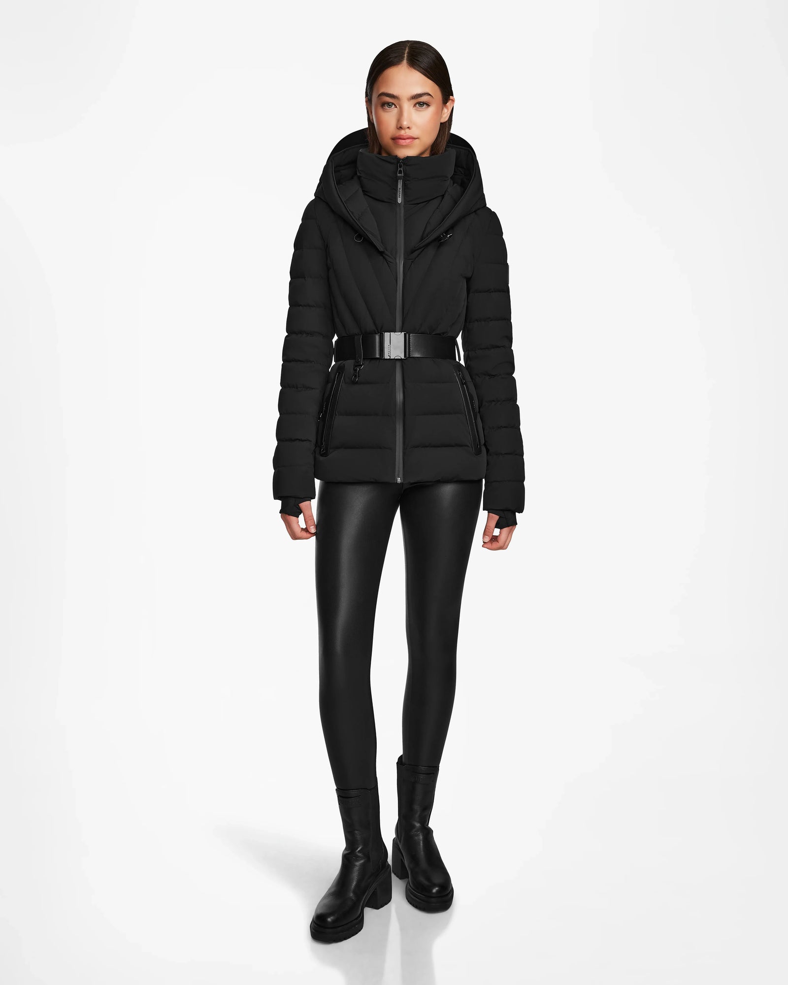 A woman faces forward wearing the Rudsak ALISA - L124506 Black quilted down puffer with vegan leather belt, black shiny leggings, and black boots. The RDS-certified jacket is showcased against a plain white background.