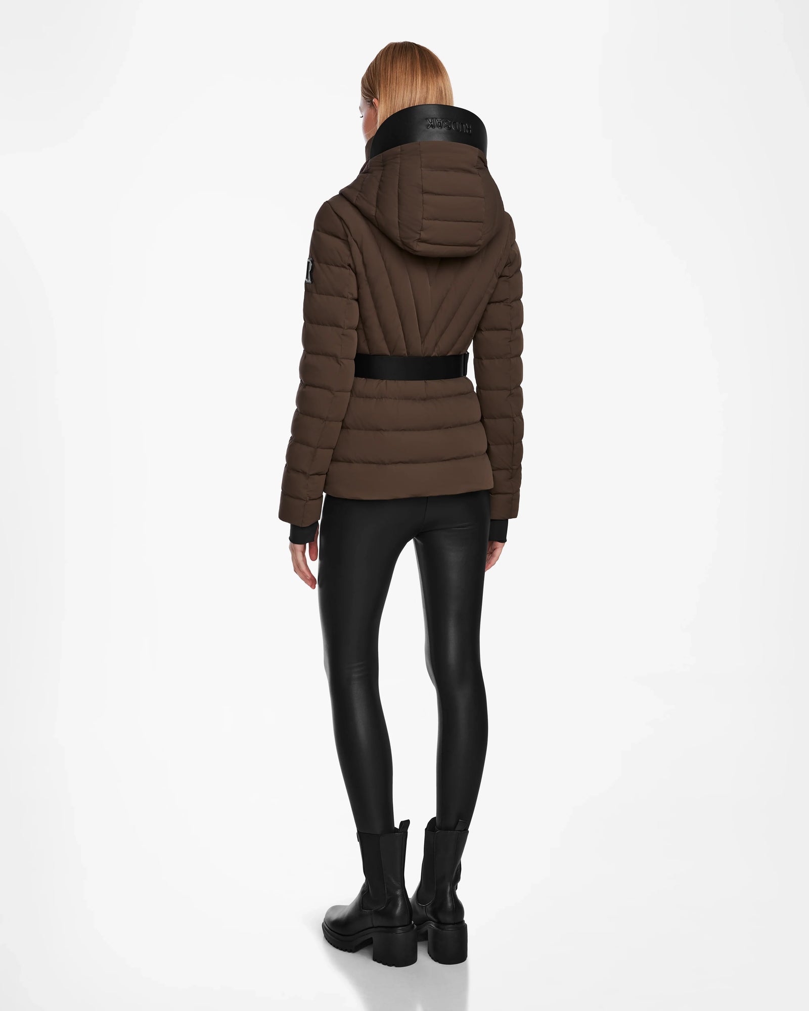 A person with long hair is seen from behind wearing the Rudsak ALISA - L124506 Espresso women’s quilted down puffer coat with hood and vegan leather belt, paired with black shiny leggings and boots, against a white background.