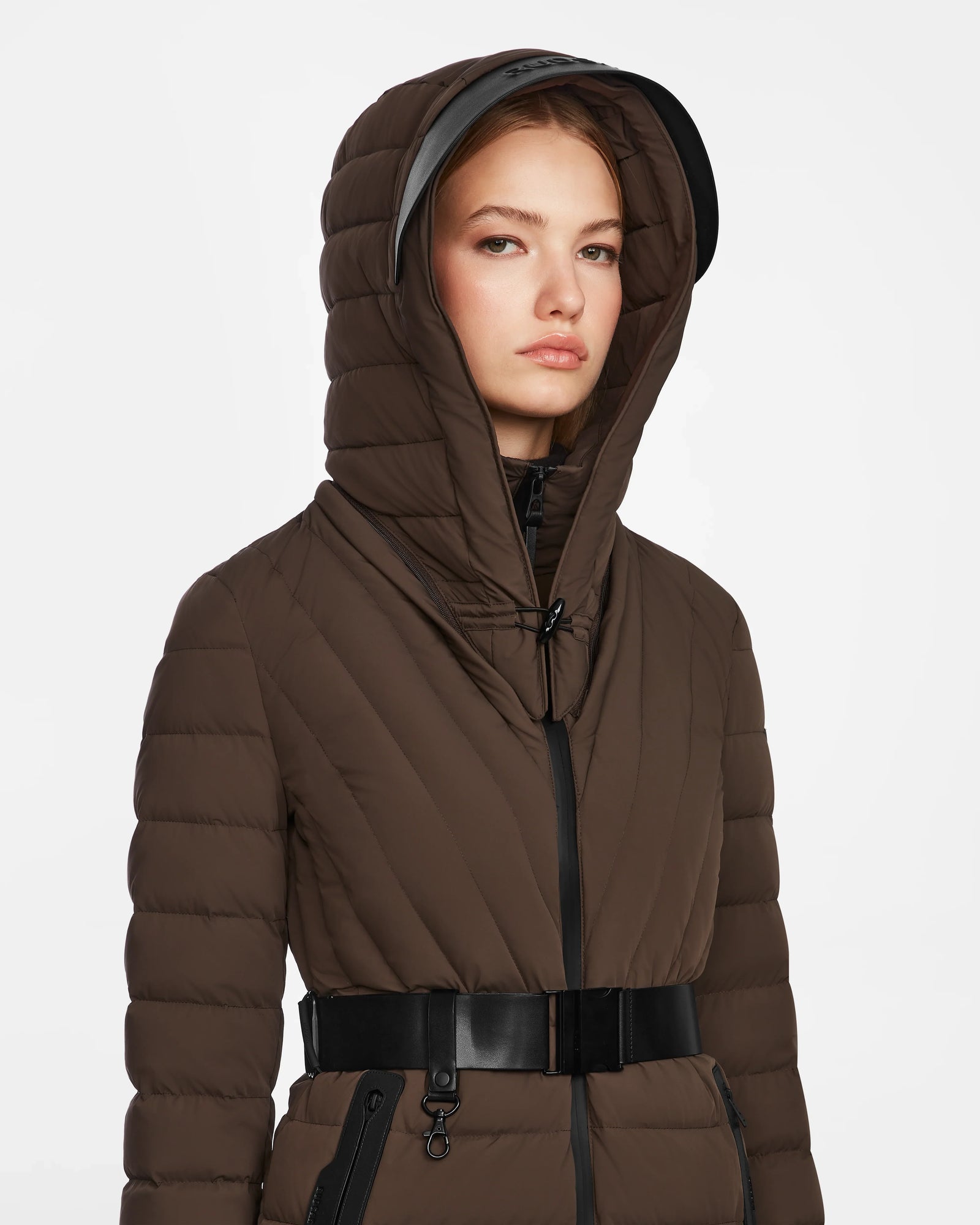 A woman wears the Rudsak ALISA - L124506 Espresso quilted down puffer winter coat with hood up and vegan leather belt, standing against a plain white background.