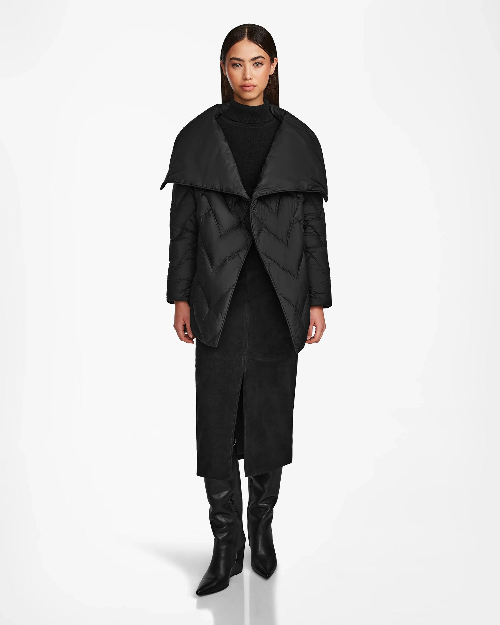 A woman faces forward against a white background, wearing the Rudsak ANNE MARIE - 8124713 Black mid-length jacket with an oversized collar, paired with a black turtleneck, long front-slit skirt, and knee-high boots.