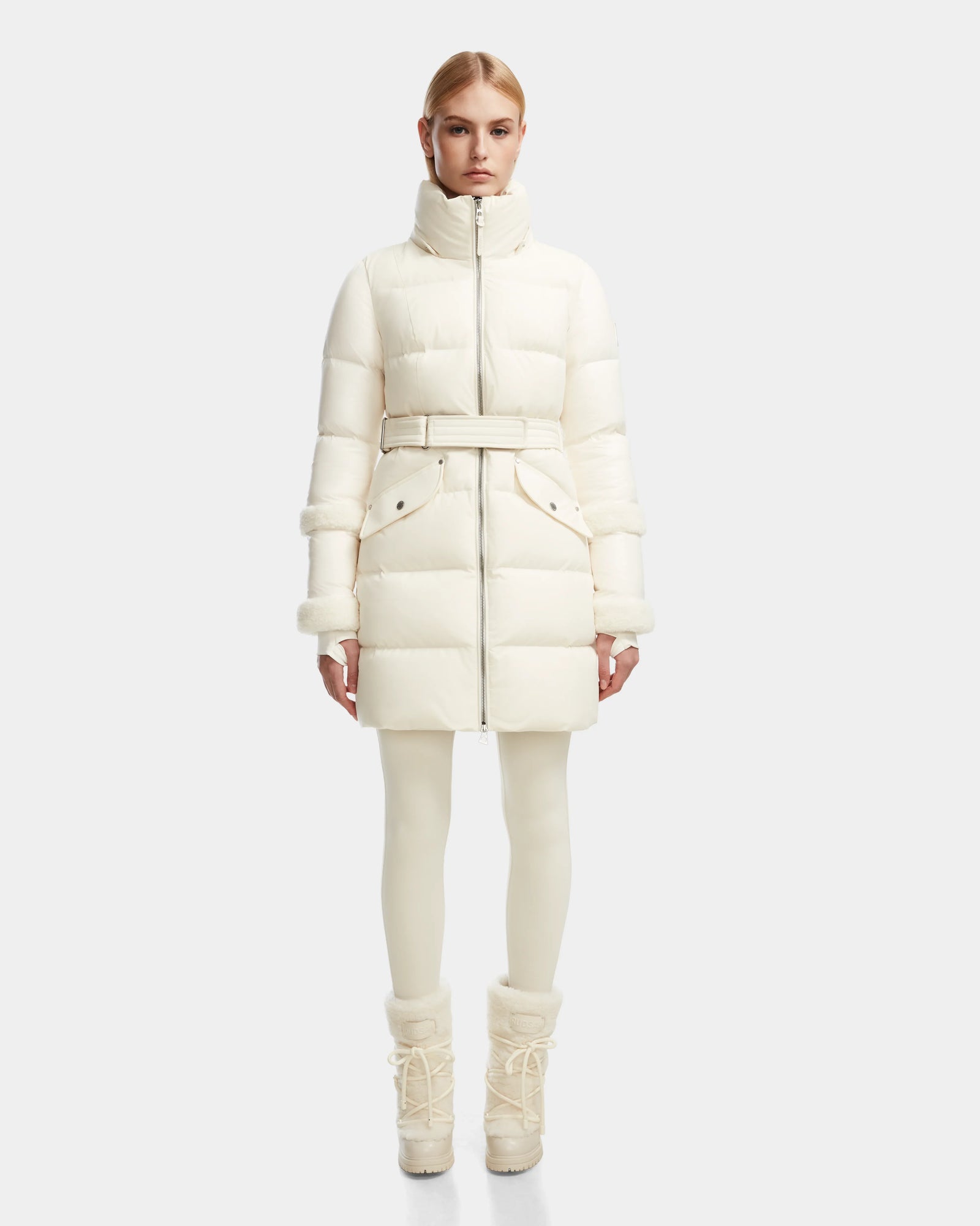 A woman faces forward wearing the Rudsak AUDREY L - 8124585 Iceland fitted, knee-length down puffer jacket with a belted waist and high collar, crafted from recycled Stormshell fabric, paired with white leggings and lace-up winter boots.