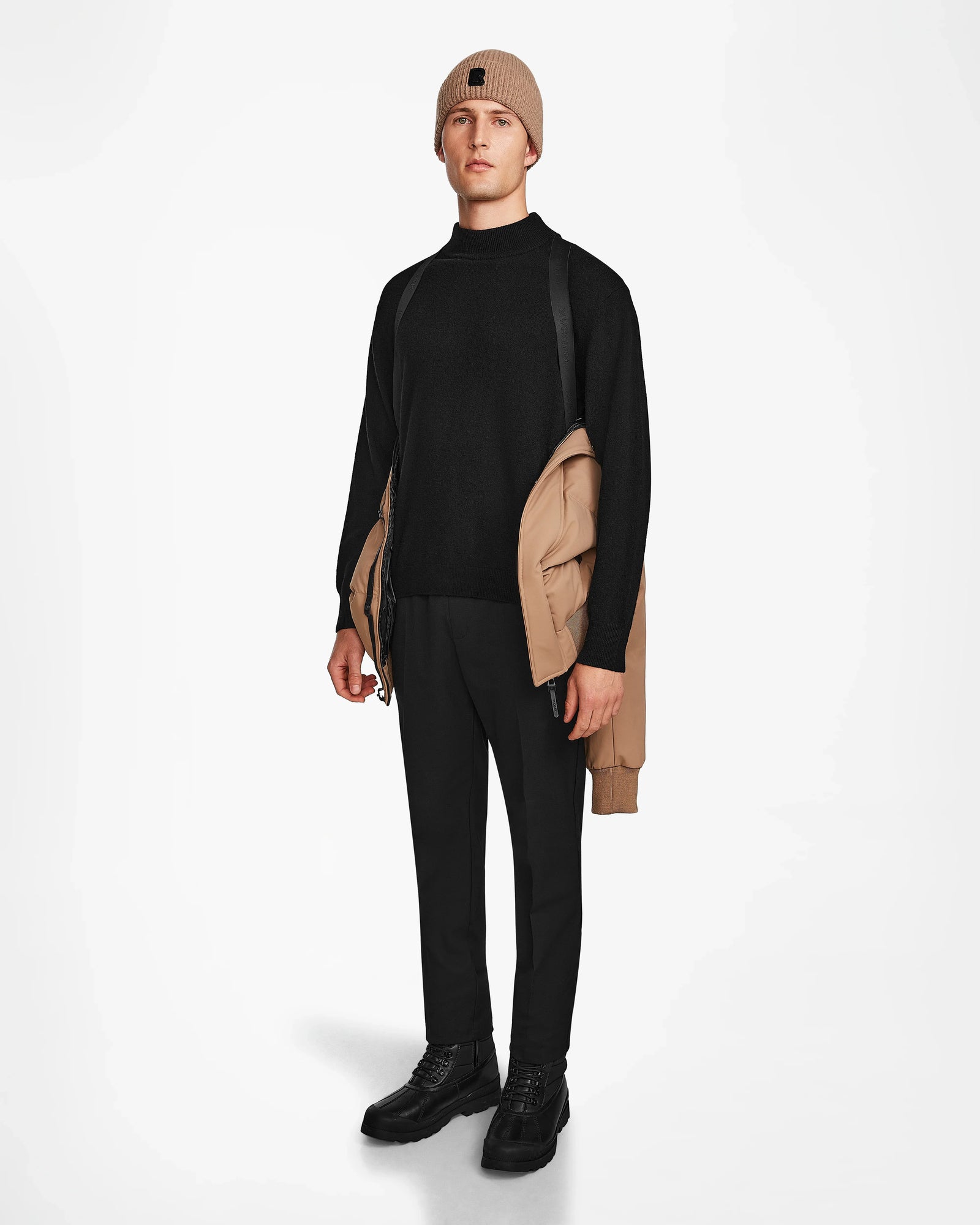 A person stands facing forward, holding the Rudsak DALLAS - M122501 Sand men's quilted down bomber over one shoulder, dressed in a beige beanie, black sweater and pants, and black shoes against a plain white background.