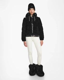 A person wearing the Rudsak DANI S - 8124537 Black bomber-style ski jacket, a black beanie, white leggings, and large black furry boots stands against a plain white background.