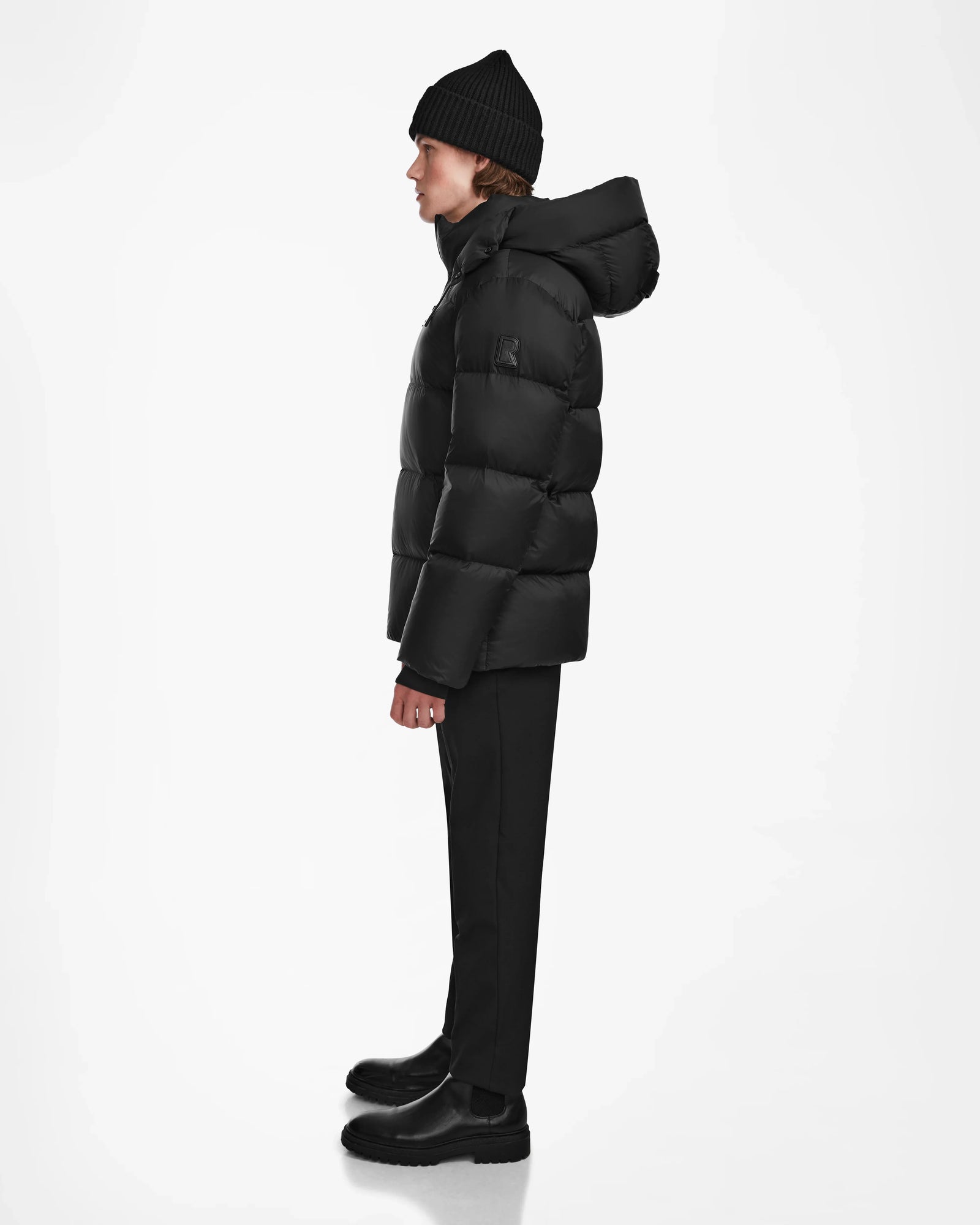 A person in profile faces left, wearing the Rudsak DANIEL M - M124510 Black hooded down puffer jacket with black pants and boots—an ideal winter outerwear choice—set against a plain white background.