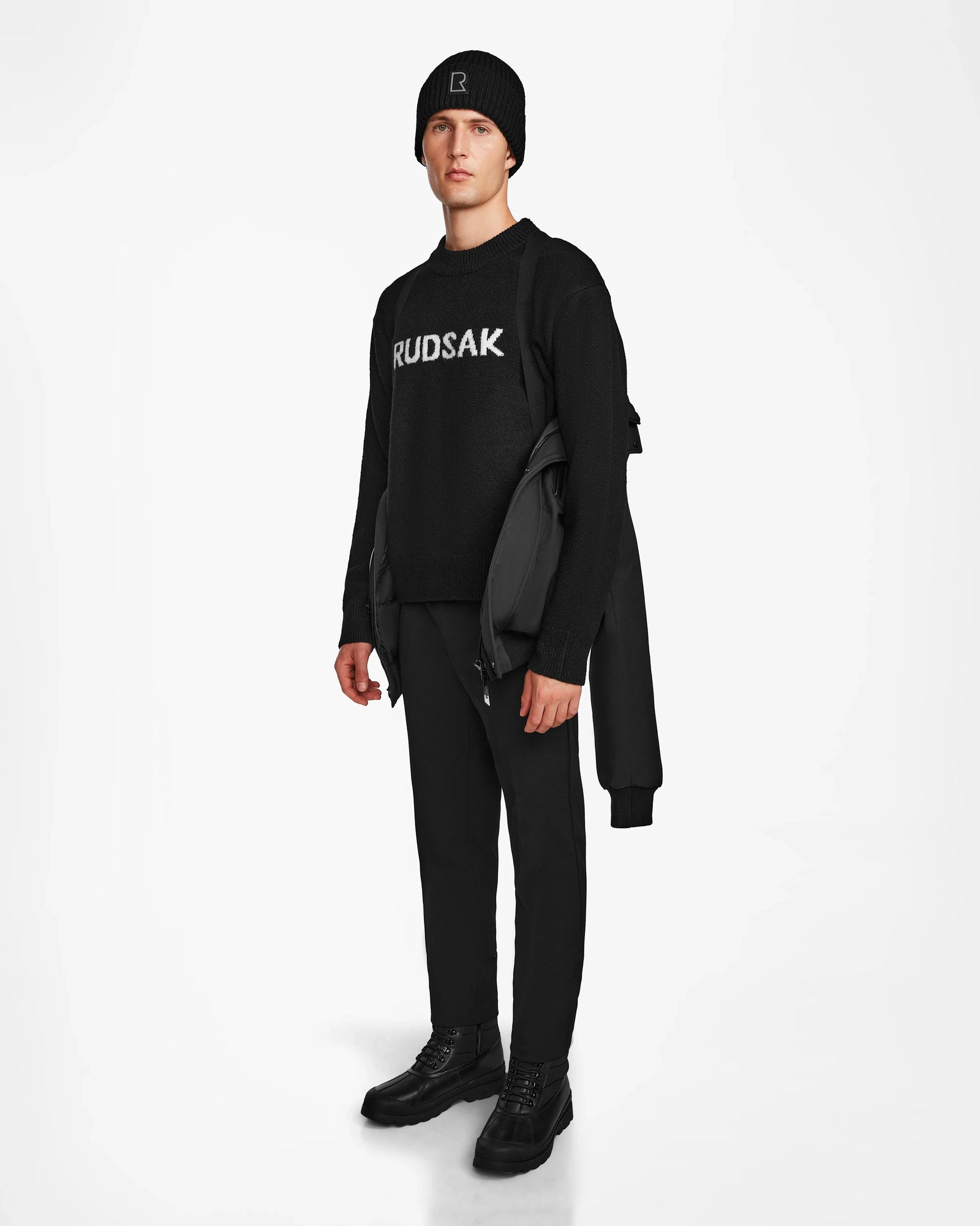 A man stands facing forward against a white background, wearing a black Rudsak sweater, black pants, and shoes. He holds the Rudsak DEACON - M123513 Black/Black mens winter coat over his left arm and wears a black beanie.