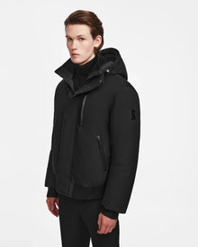 A young man with light skin and brown hair models the Rudsak DEACON - M123513 Black/Natural bomber-style down puffer with RDS-certified down, standing against a plain white background and looking slightly to the side.