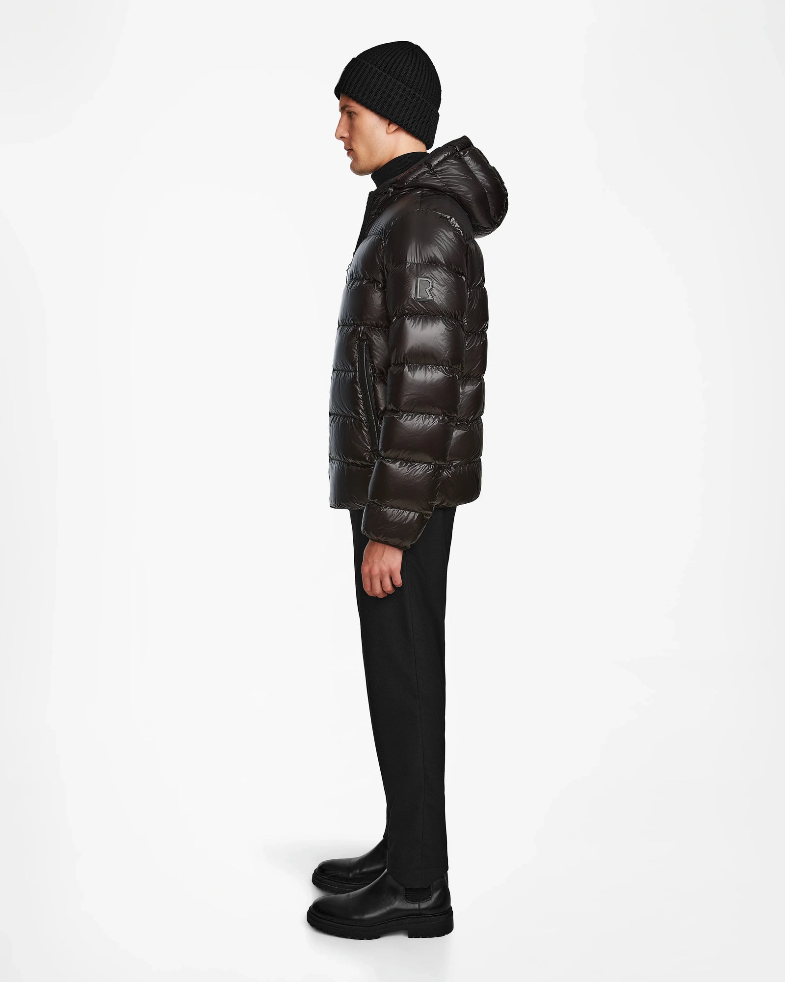 A man stands in profile against a white background, wearing the Rudsak DEMIAN S - M123508 Black men’s down puffer jacket, paired with black pants, black boots, and a black knit beanie.