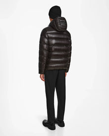 A person stands facing away in a Rudsak DEMIAN S - M123508 Black mens shiny down puffer jacket with hood, water- and wind-resistant, paired with black pants, shoes, and a knit beanie against a plain white background.