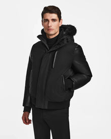 A man in the Rudsak DERON - M123517 BLACK mens bomber jacket, featuring a fur-lined hood, stands against a white background with one hand in his pocket. This stylish weather-resistant piece is perfect for chilly days.