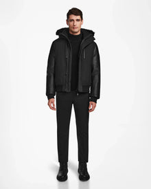 A man stands facing forward in the Rudsak DERON - M123517 BLACK mens bomber jacket, featuring leather sleeves and weather resistance, paired with black pants, sweater, and boots against a plain white background.