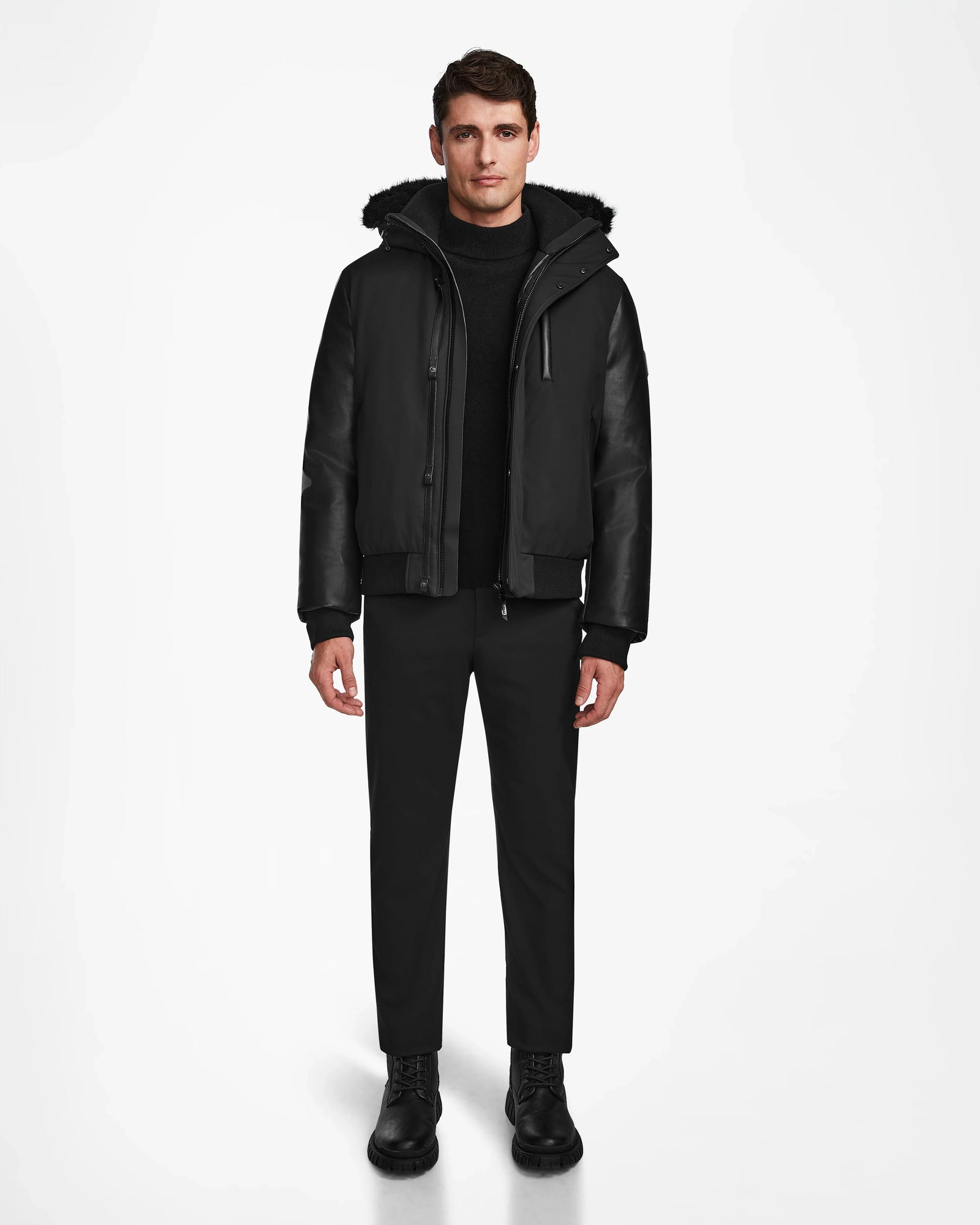 A man stands facing forward in the Rudsak DERON - M123517 BLACK mens bomber jacket, featuring leather sleeves and weather resistance, paired with black pants, sweater, and boots against a plain white background.