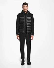 A man faces forward, wearing the Rudsak DURIN S - M123518 Black mens hooded jacket with horizontal quilting and lightweight down insulation, paired with black pants and black boots, set against a plain white background.