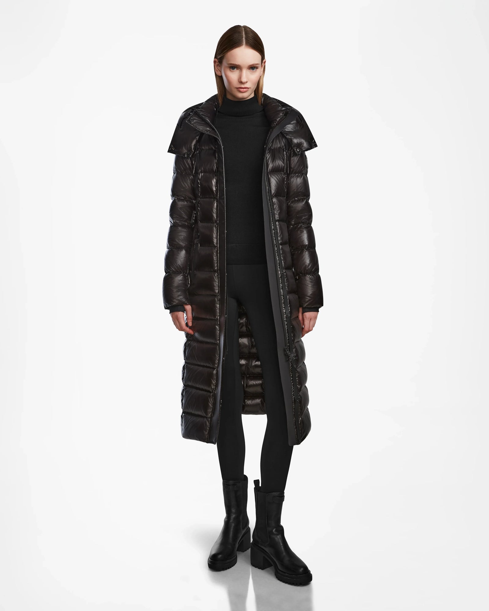 A woman faces forward in the Rudsak FRIDA L-S - L124525 Black belted puffer coat, styled over a black turtleneck, leggings, and boots—an effortlessly chic winter look. The background is plain white.