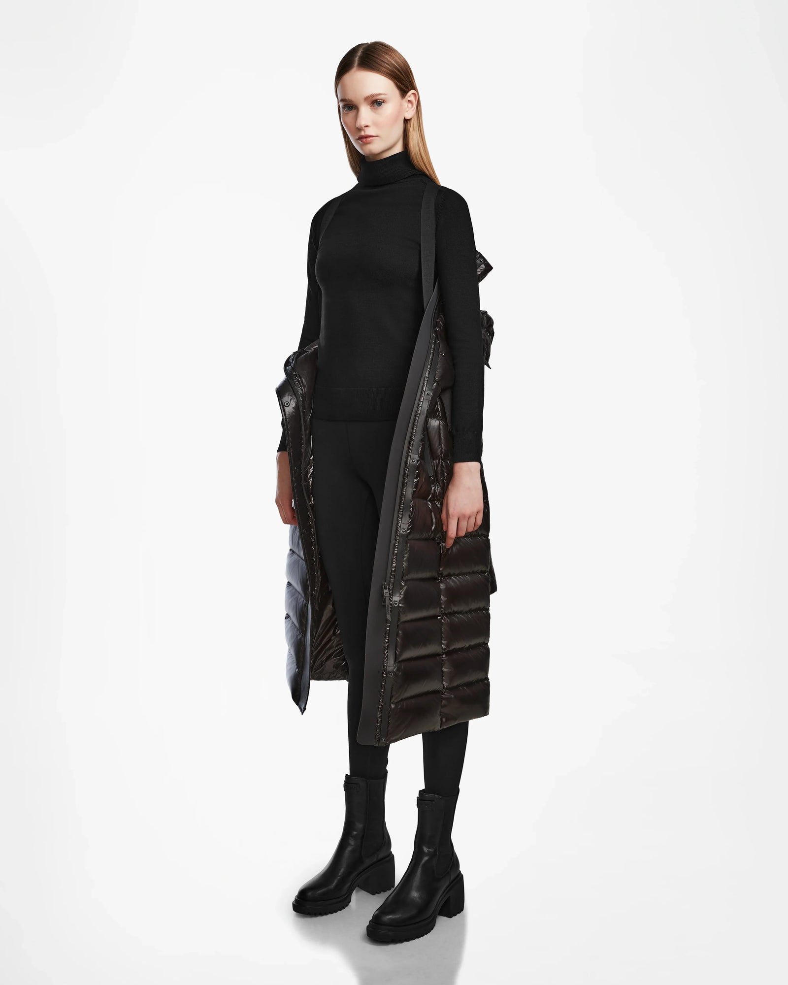 A woman stands against a white background wearing the Rudsak FRIDA L-S - L124525 Black quilted maxi down puffer over a black turtleneck, pants, and boots. Her straight hair is down and her expression is neutral—perfect for any winter wardrobe.