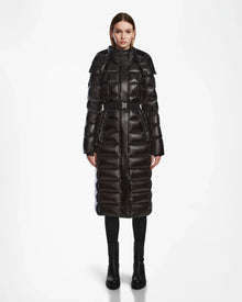A woman faces forward, modeling the Rudsak FRIDA L-S - L124525 Black, a women's quilted maxi down puffer with high collar and belt, paired with black boots. The plain white background highlights this winter essential.