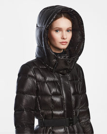A woman models the Rudsak FRIDA L-S - L124525 Black belted quilted puffer with a large hood against a white background. The shiny black coat adds chic style to any winter wardrobe as she gazes to the side with a neutral expression.
