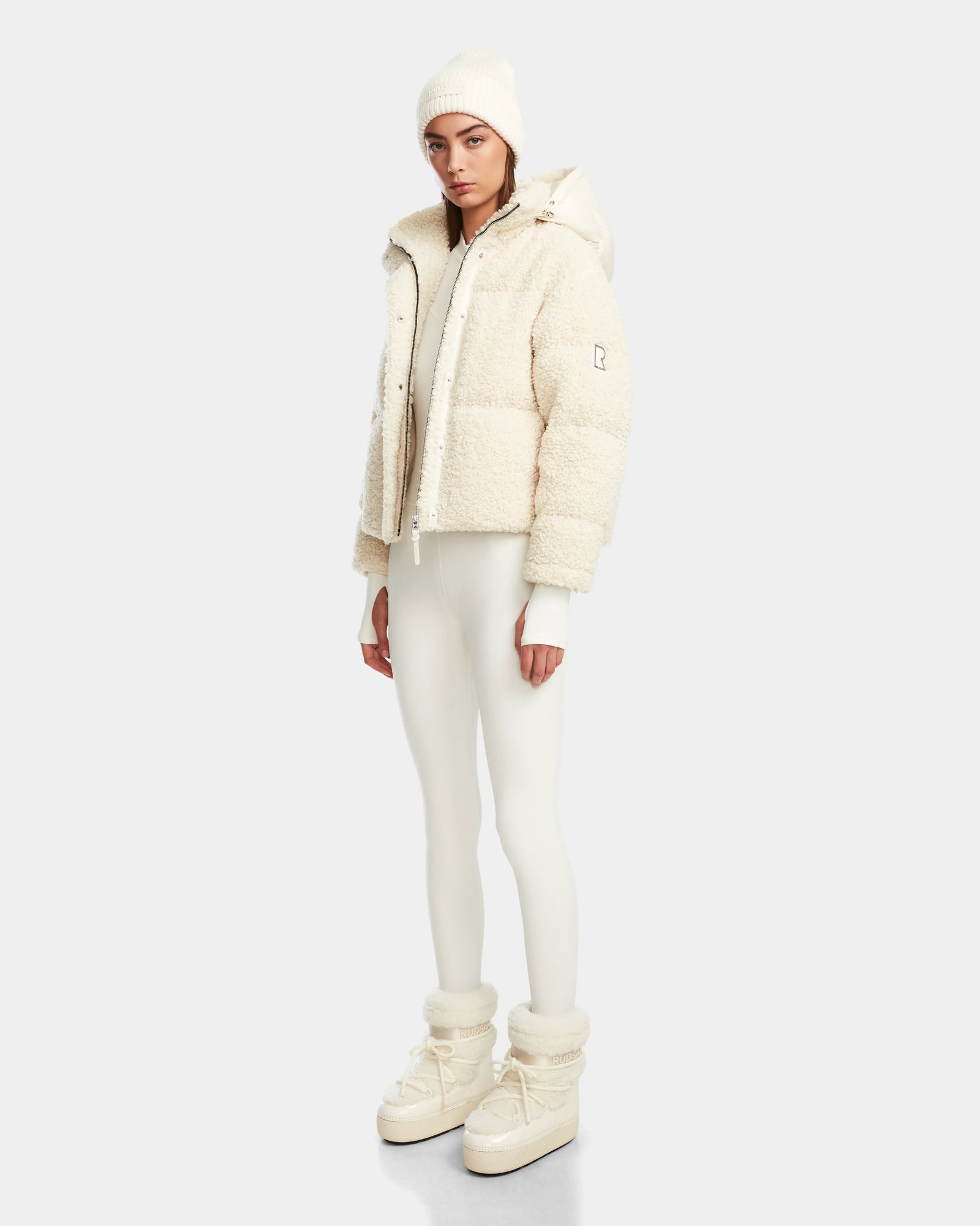A person stands against a plain background, dressed in the Rudsak IKSA - 8124706 Iceland women’s teddy down puffer, styled with a cream beanie, fitted leggings, and high-top boots for an effortlessly chic off-white winter look.