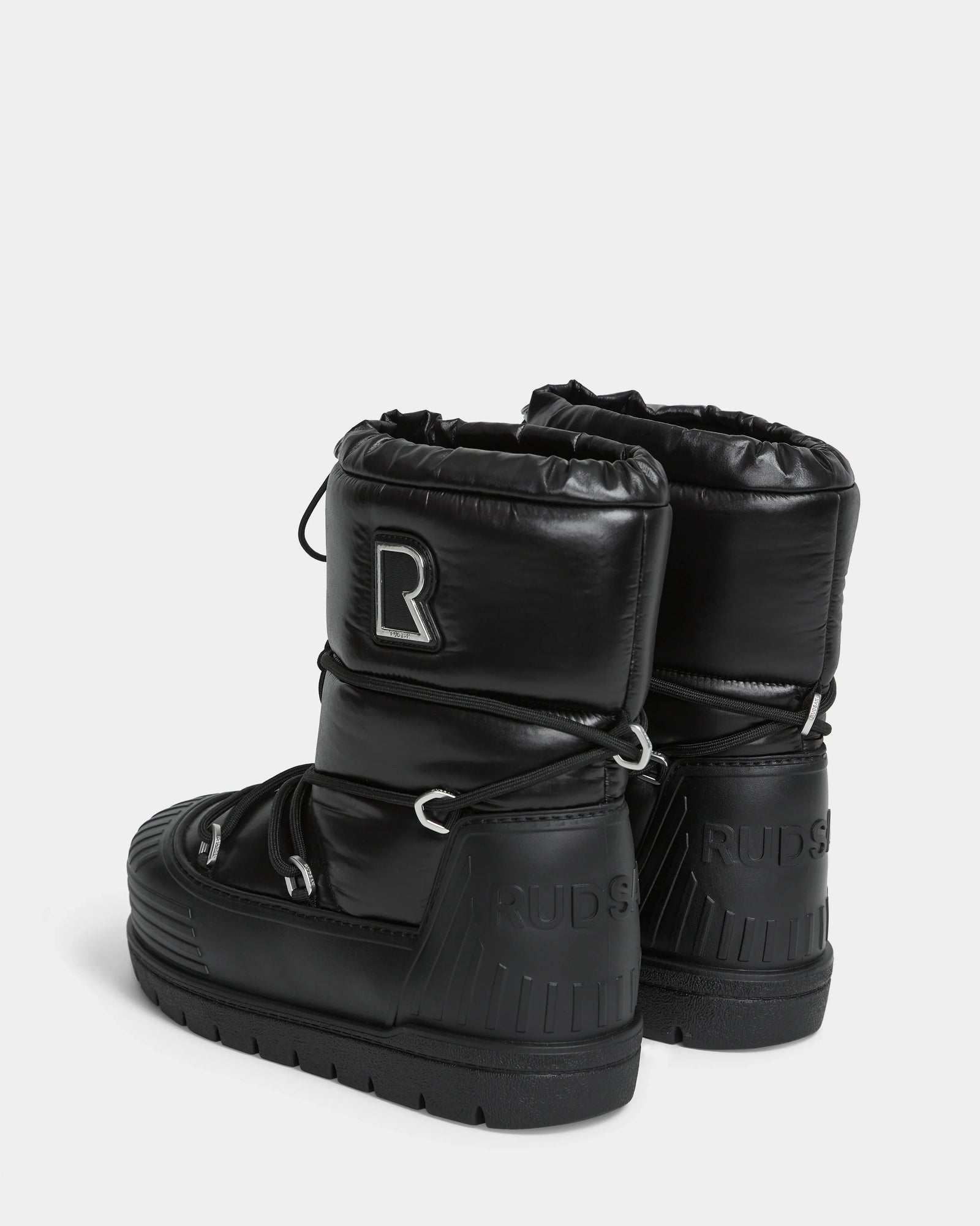 The KIKA - L224506 Black by Rudsak are shiny black winter boots with chunky rubber soles, a large R patch on one boot, Rudsak embossed on the heels, drawstring closures, and stylish rubber detailing.