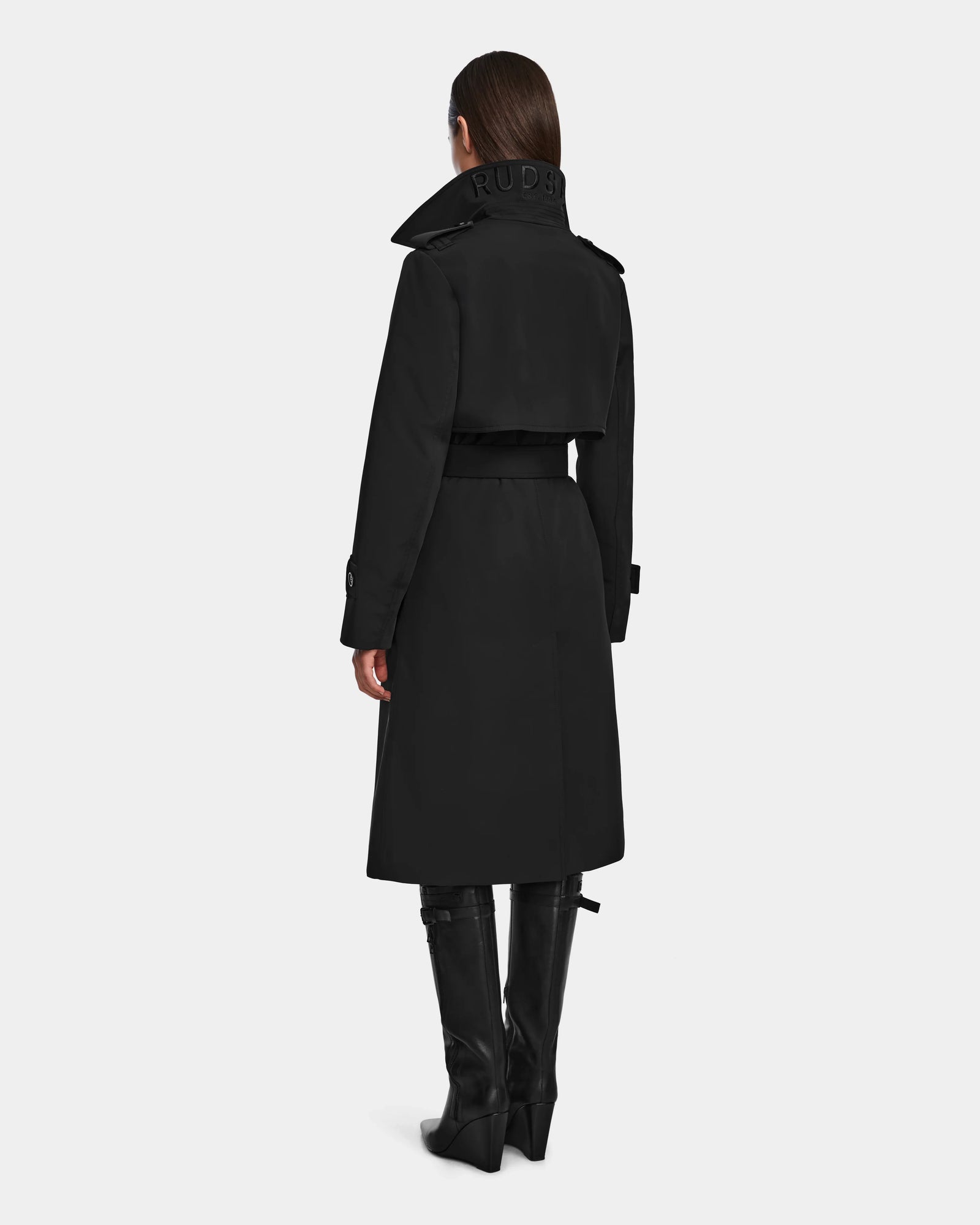 A woman stands facing away, wearing the Rudsak KINDA - 8124690 Black womens trench coat with leather detailing, a high collar, embossed lettering at the back, waist belt, and knee-high black boots.