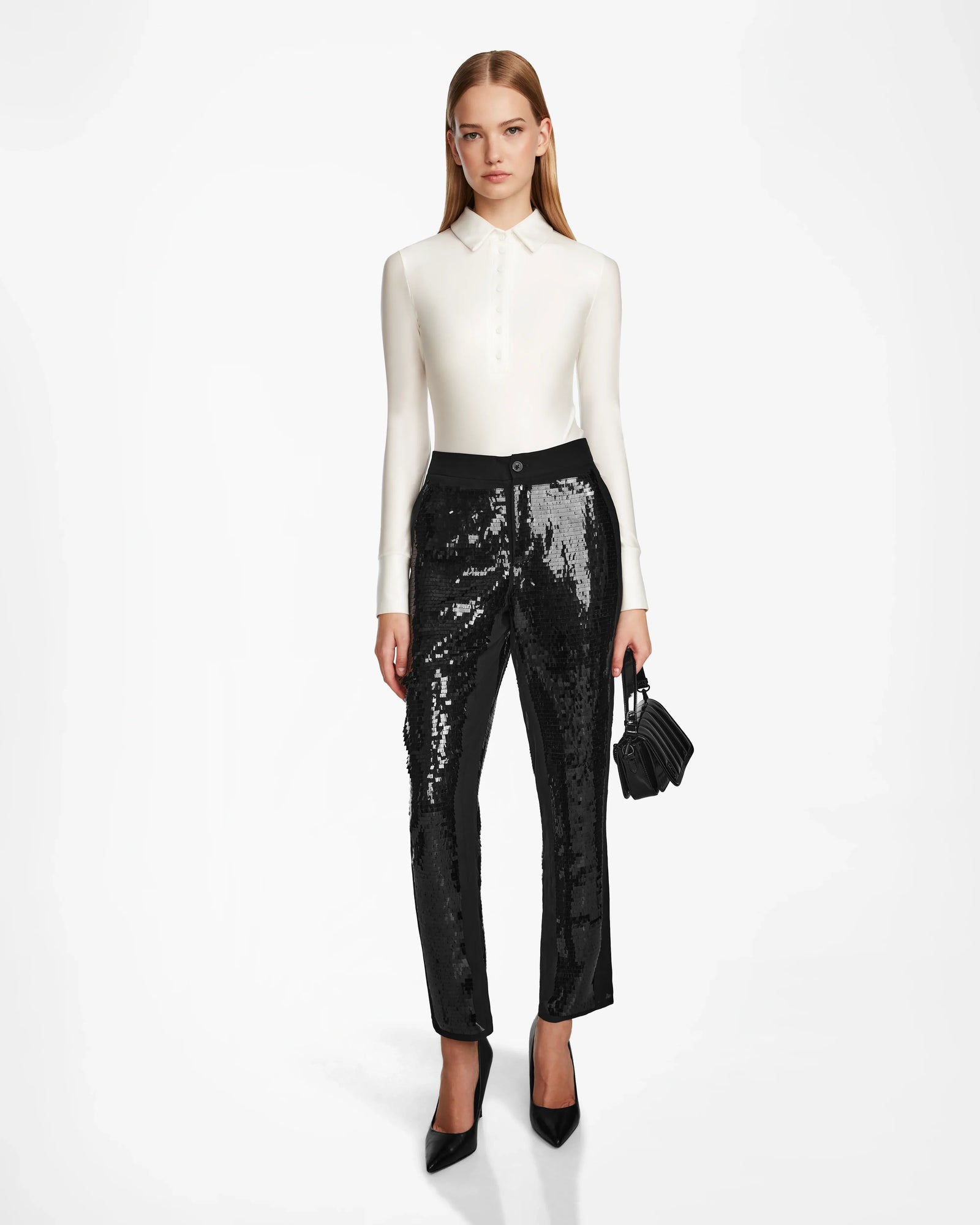 A woman stands against a white background wearing a fitted white long-sleeve blouse, festive sequin pants, black pointed-toe heels, and holding the Rudsak LEAR - 8524553 Black handbag.