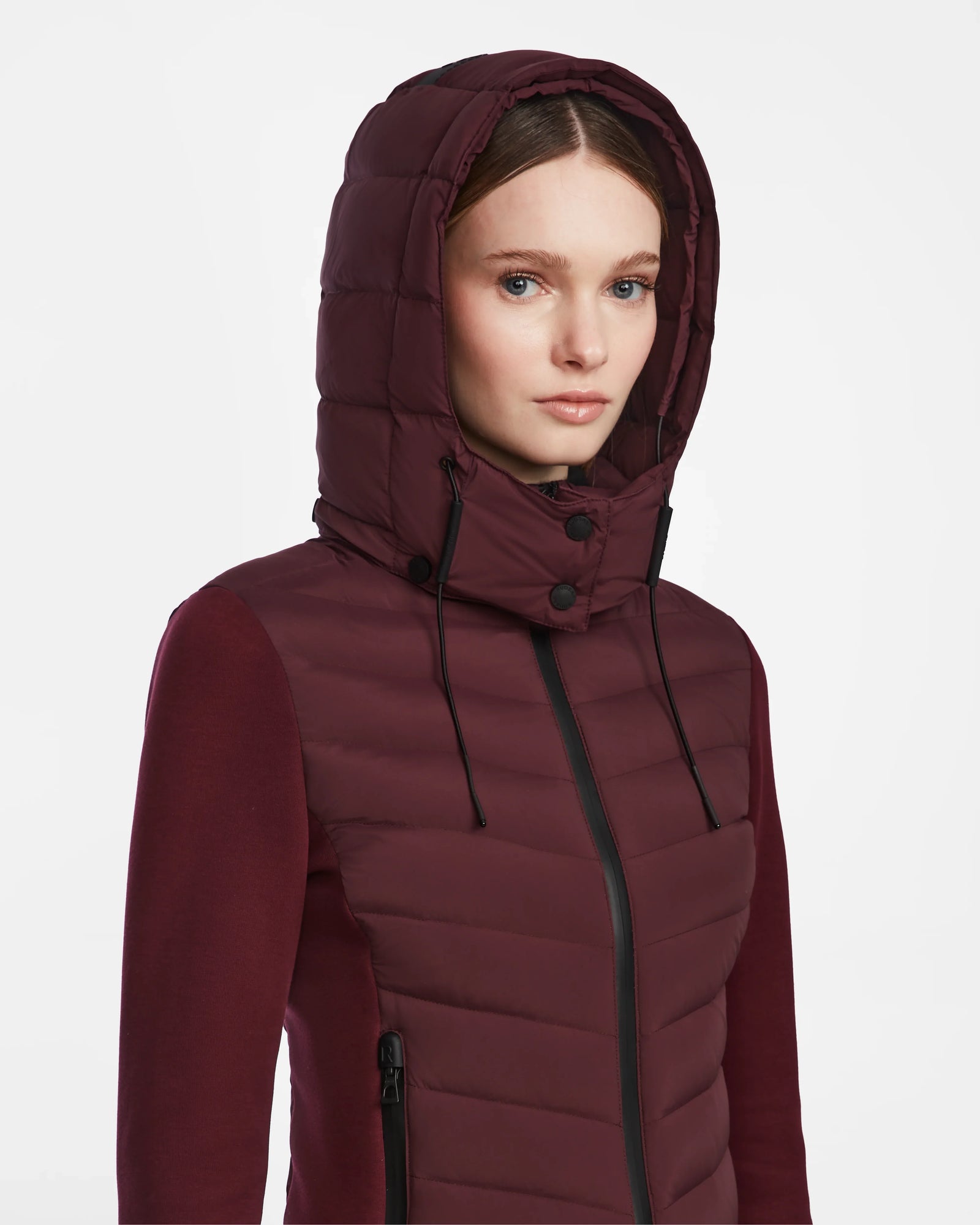 A woman models the Rudsak MALYA M - L123547 Wine jacket, a maroon down quilted style with hood, black zippers, and drawstrings—perfect as a cozy lightweight jacket for women.