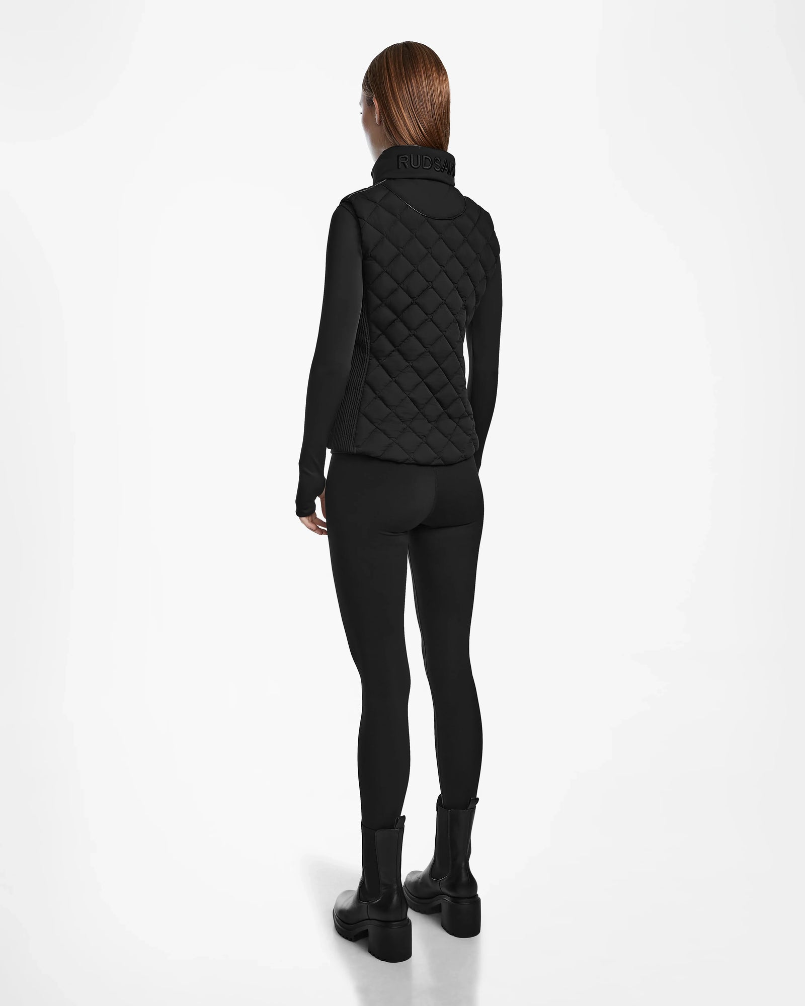 A person faces away, wearing the Rudsak MATILDA - L124504 Black quilted women’s light vest over a black long-sleeve top, leggings, and boots—ideal for transitional weather—against a plain white background.