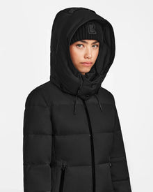 A person wears the Rudsak MIRIAM - L122523 BLACK womens down puffer with a weather-resistant shell and a black knit beanie featuring an R, looking forward against a plain white background.