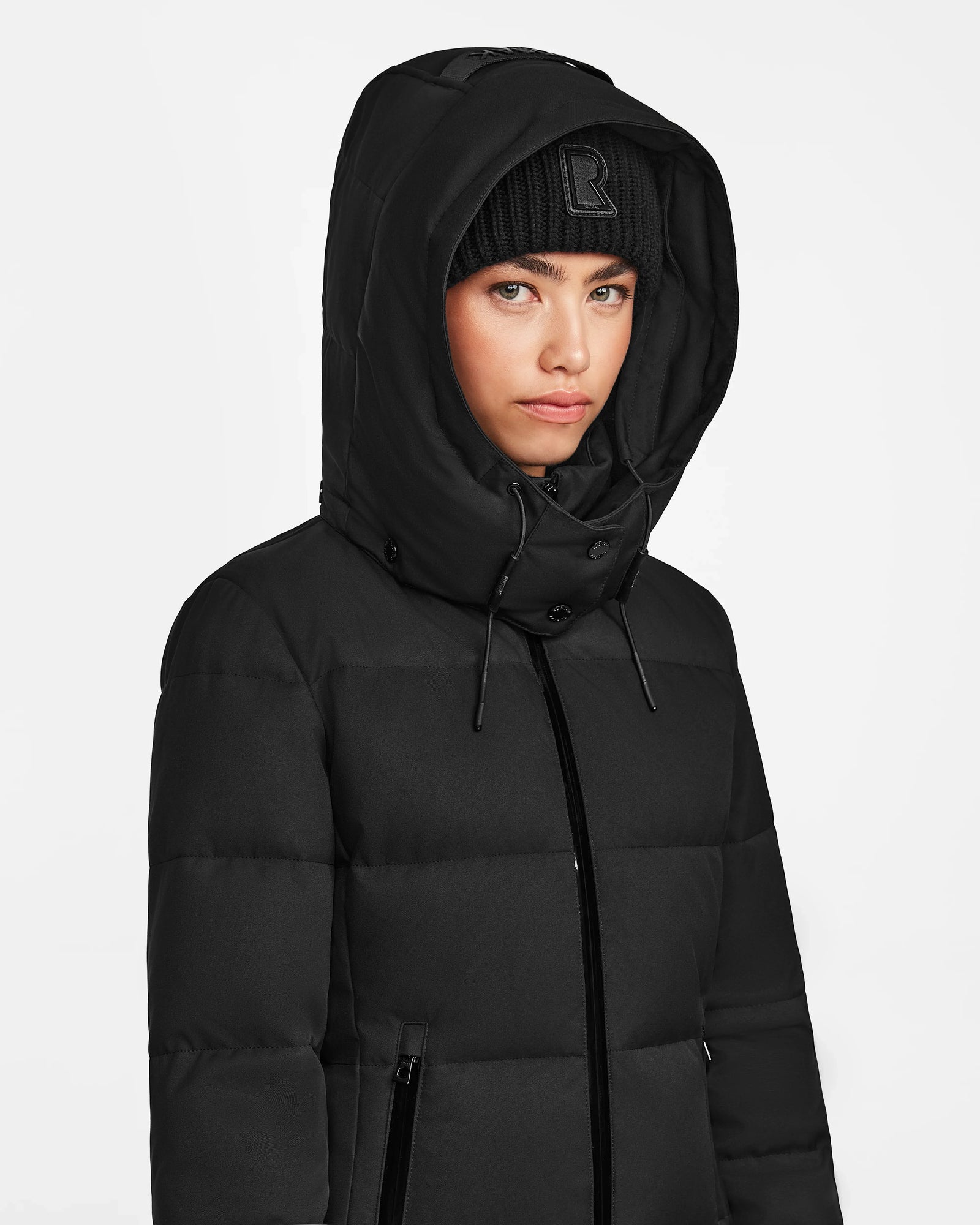 A person wears the Rudsak MIRIAM - L122523 BLACK womens down puffer with a weather-resistant shell and a black knit beanie featuring an R, looking forward against a plain white background.