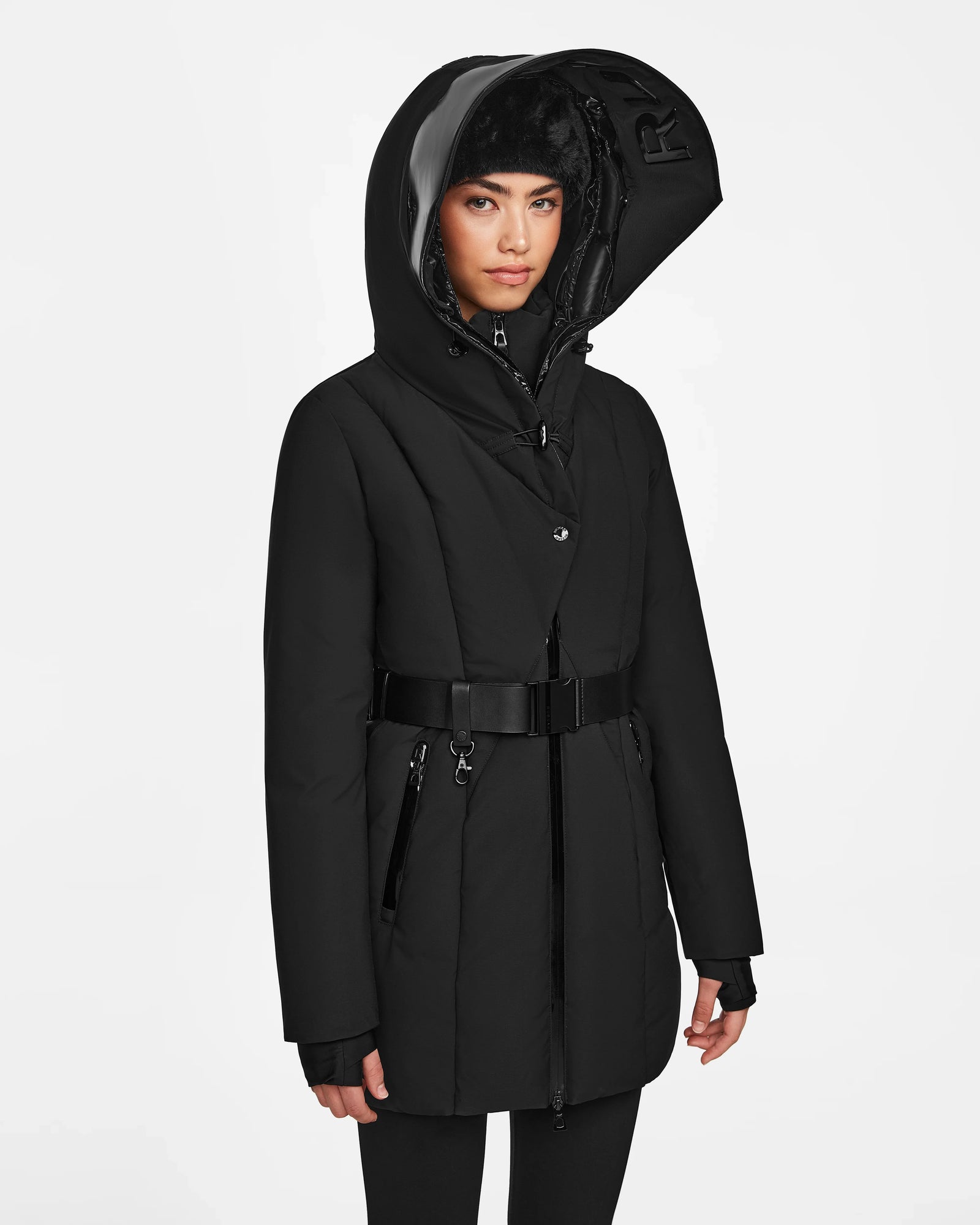 A woman models the Rudsak MODA M - 8124555 Black, a belted black winter coat with a hood, high collar, and zipper, featuring sustainable design elements, against a plain white background.