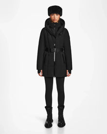 A person stands facing forward in the Rudsak MODA M - 8124555 Black winter coat with belt, black leggings, boots, and a fur hat against a white background—showcasing timeless style and sustainable design.