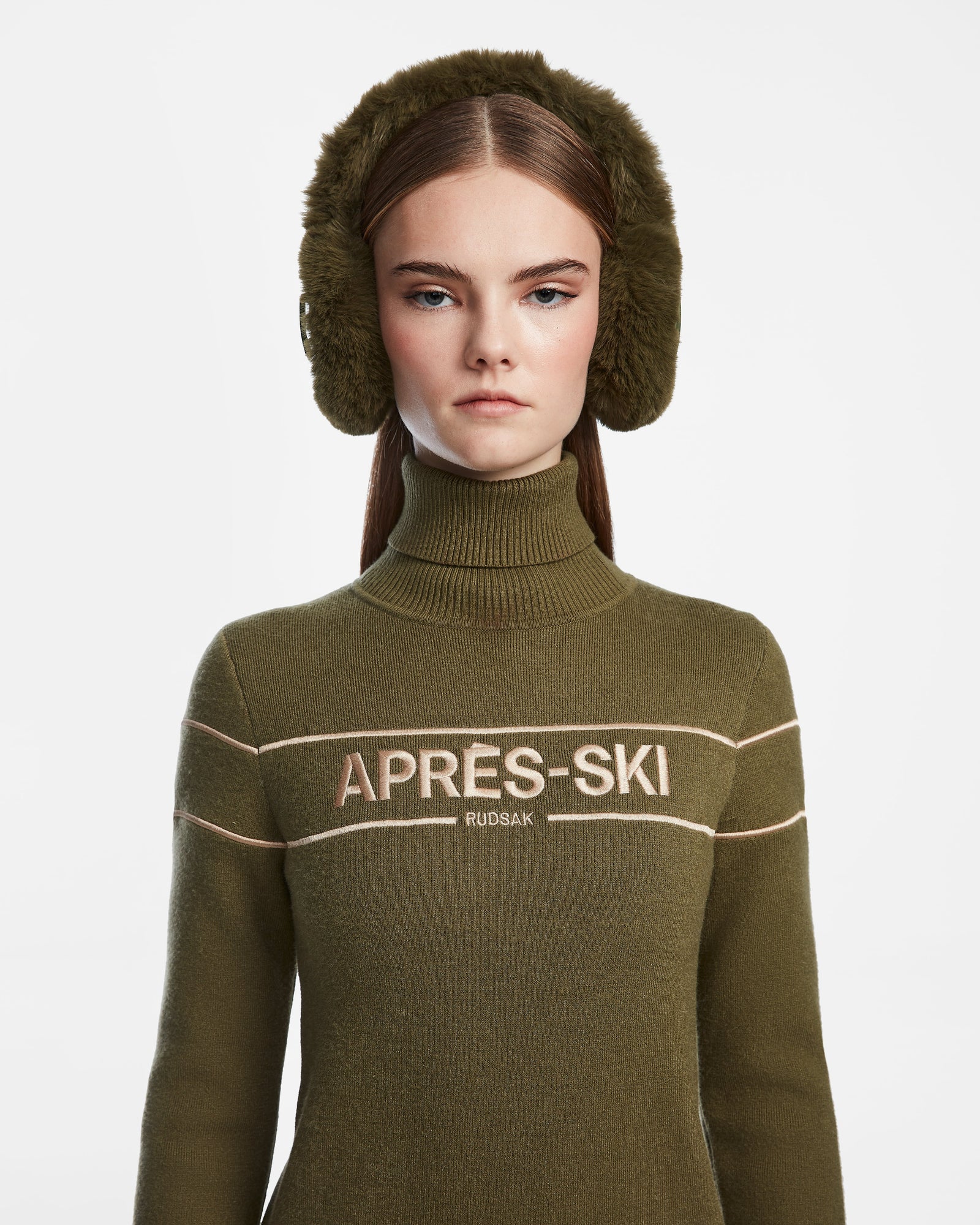 A woman with straight brown hair models large Rudsak MUFF - 8422503 Loden Green earmuffs and an olive green turtleneck with APRÈS-SKI RUDSAK on the front, standing against a white background—a chic winter accessory look.