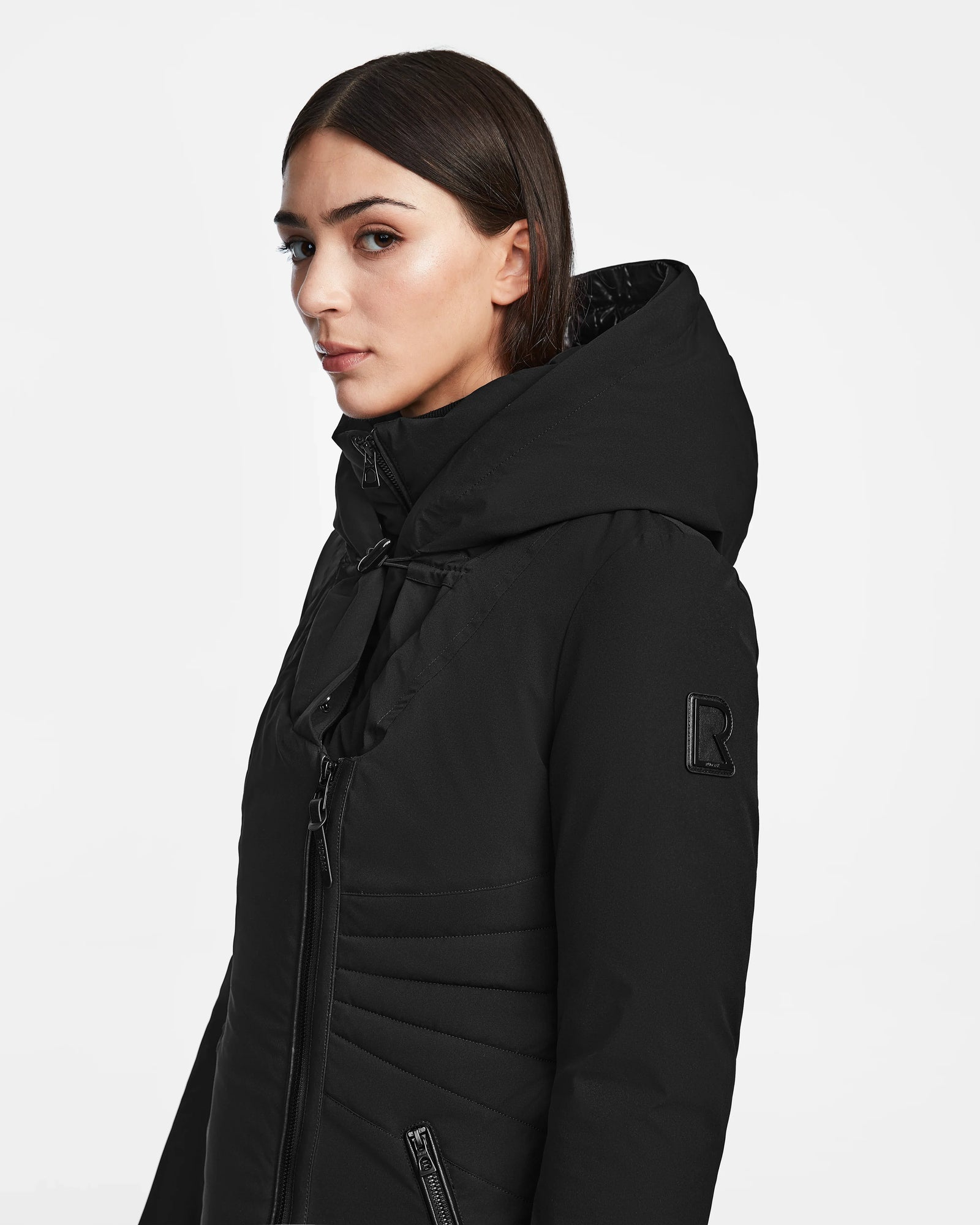 A woman with straight brown hair models the Rudsak MUSA - L124534 Black women’s long down puffer, looking slightly to the side against a plain white background.