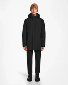 A person faces forward against a plain white background, wearing the Rudsak NATAN - M124519 Black mens parka, paired with black pants, black boots, and a brown knit beanie.