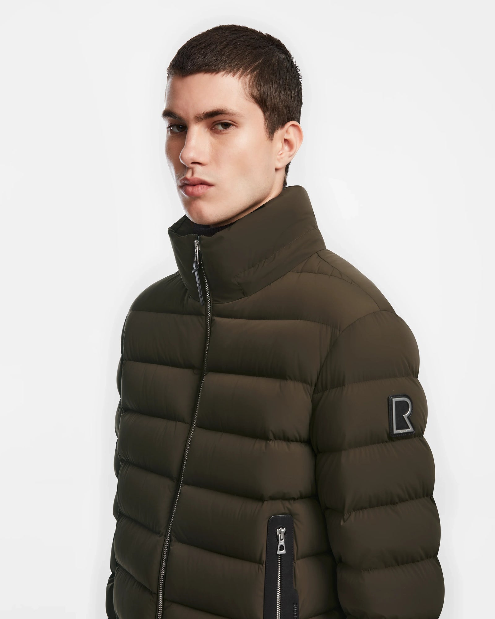 A young man with short dark hair models the Rudsak NAVAN - 6124551 Army olive green quilted puffer coat with a high collar and black sleeve patch, standing against a plain white background.