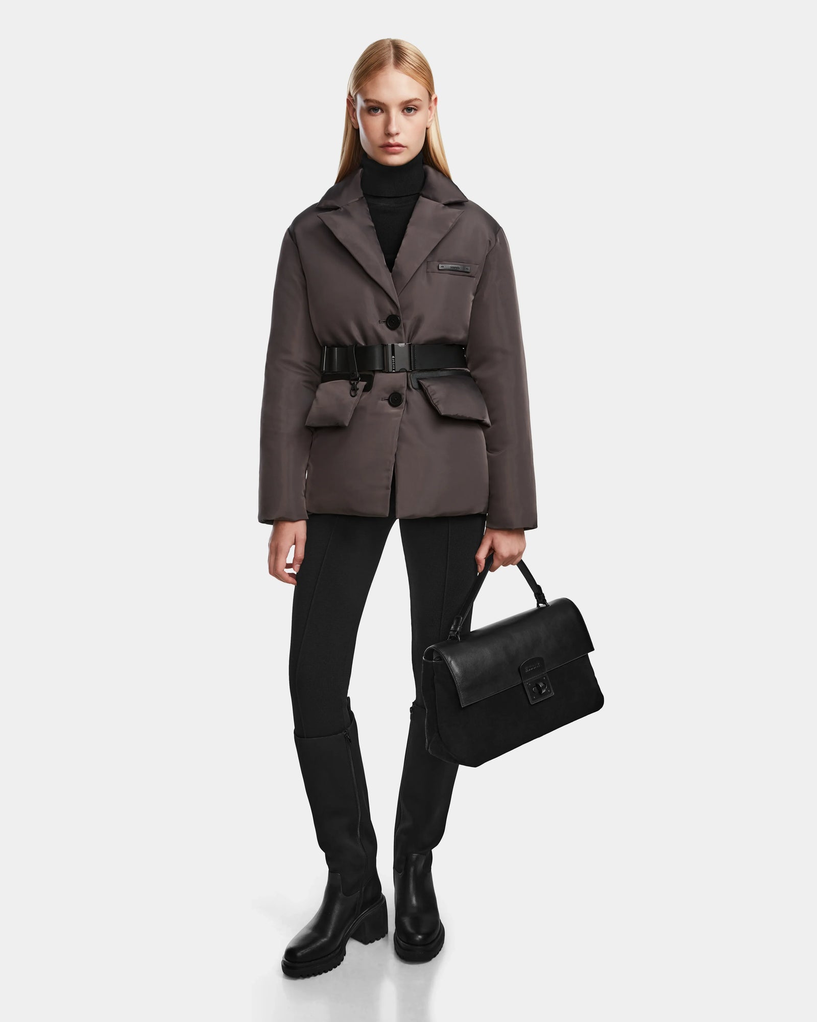 A woman stands against a white background wearing the Rudsak PAOLA - 8124547 Grey down blazer, paired with a black turtleneck, black pants, knee-high black boots, and holding a black handbag at her side.