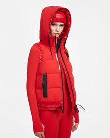 A person wears the RAQUEL M - 8124538 Flame Red ski vest by Rudsak over a red fitted jacket, red leggings, and a red beanie, styled with black zipper and drawstring accents for a high-performance look against a white background.