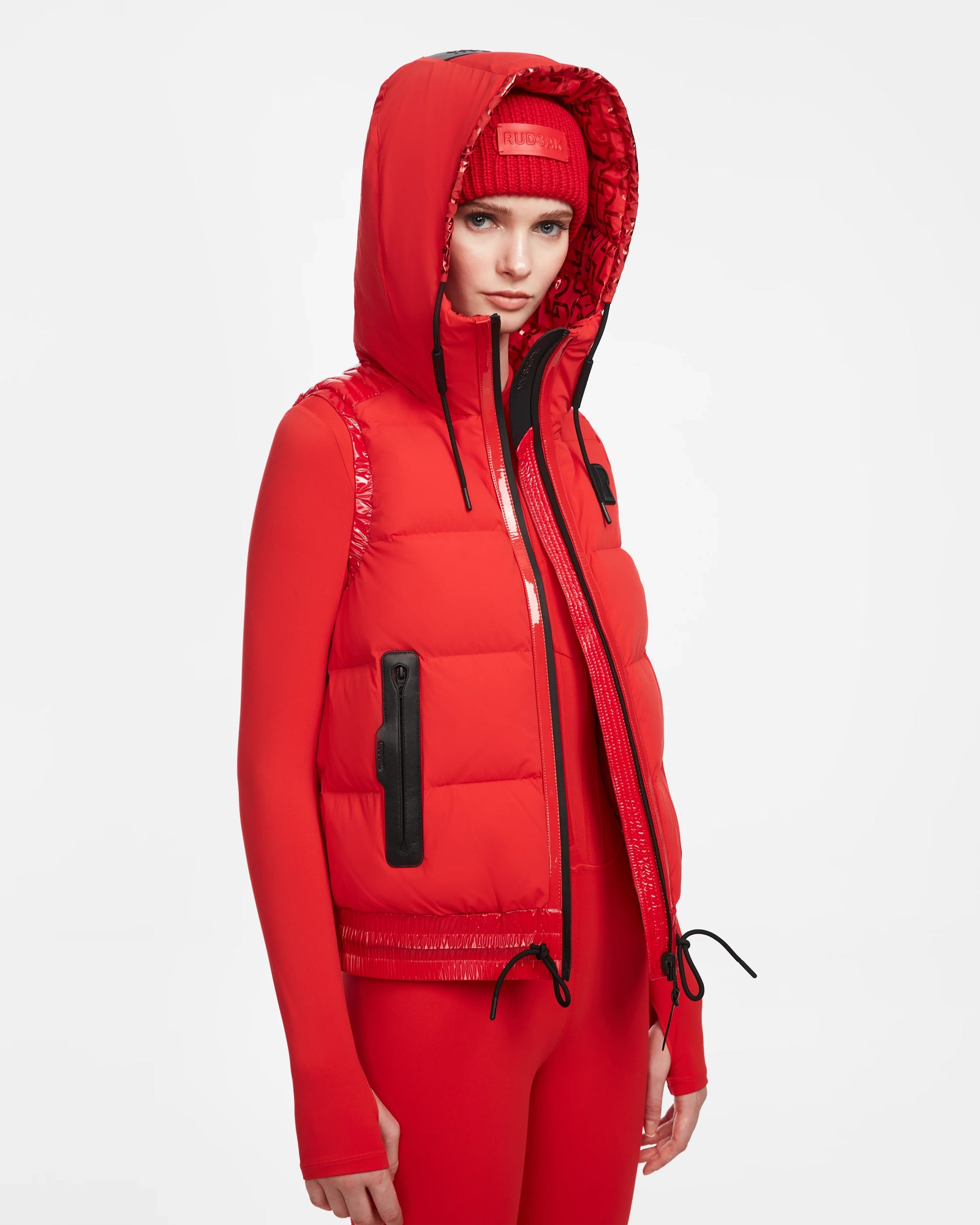 A person wears the RAQUEL M - 8124538 Flame Red ski vest by Rudsak over a red fitted jacket, red leggings, and a red beanie, styled with black zipper and drawstring accents for a high-performance look against a white background.