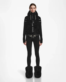 A person stands facing forward in the Rudsak RAQUEL S - 8124526 Black ski vest, black top, fitted pants, and large black fuzzy boots—ready for winter mountain adventures against a plain white background.