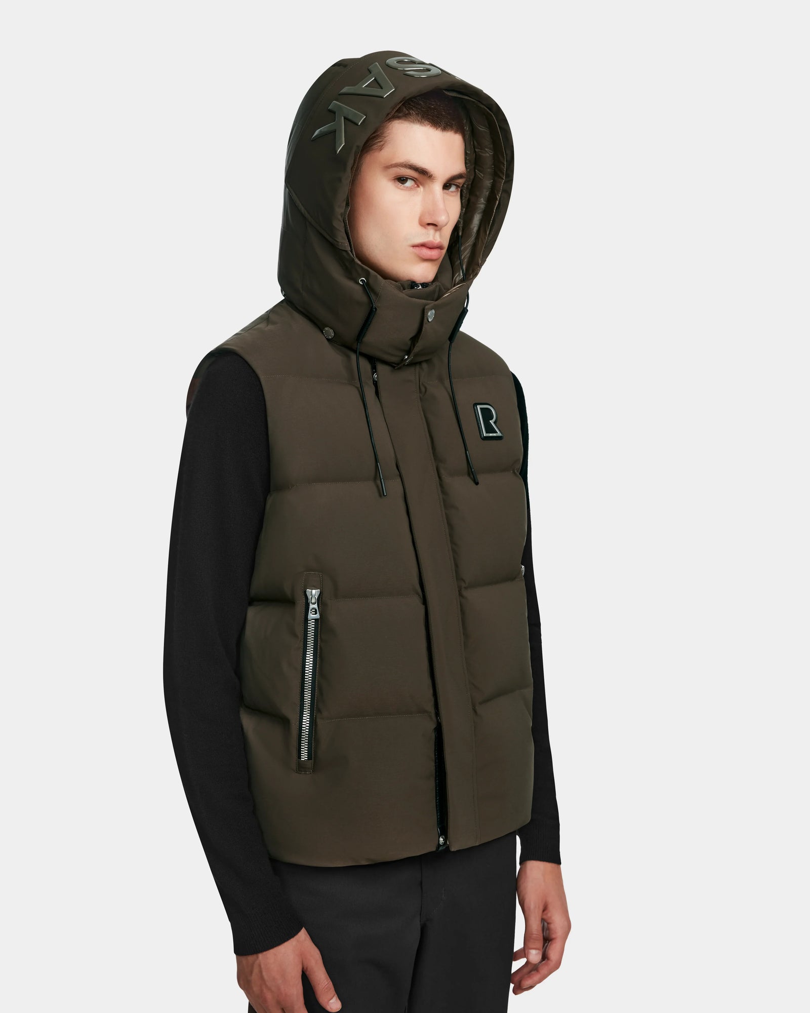 A young man wears the Rudsak REN M - 9123512 Army M, a weather-resistant dark green down puffer vest with an R logo on the chest, zipper front, and pockets, paired with black long sleeves and pants.